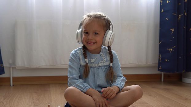 Child Listening Music In Big White Headphones. Happy Little Preschool Toothless Girl 5 Years Old Looking At Camera In Leaving Room Inside. Kid Smiles Laughs Shows Tongue At Home. Childhood, Education.