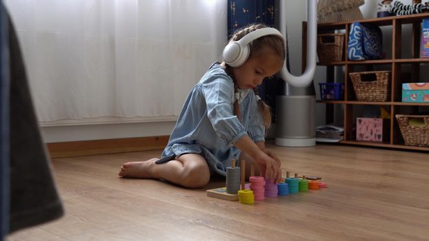 Happy Little Preschool Toothless Girl Playing With Colored Wooden Toy. Kids Learn To Count By Playing Teaches Numbers At Home. Child Listening To Music In Big White Headphones. Childhood, Education.