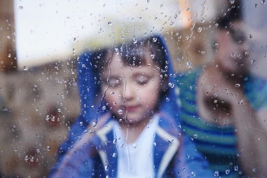 A little boy looking downwards behind a rain-streaked window while his mother sits in the background.