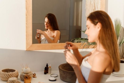 Facial skin care. Attractive woman using hyaluronic face serum, moisturizing and skin care, standing near mirror in modern indoor bathroom. Daily beauty routine