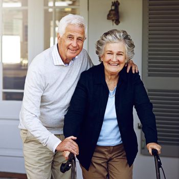 Portrait of a smiling senior woman using a walker with her husband beside her outside their home.