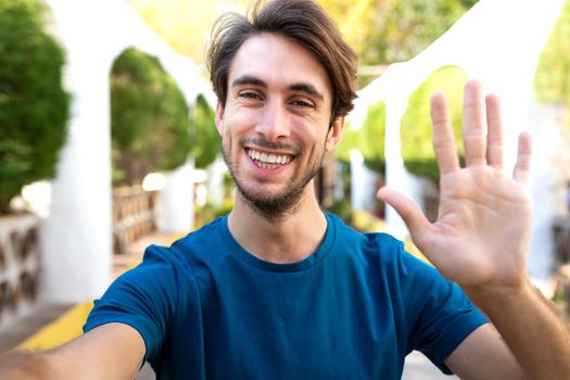 Young caucasian man wave hello on a video call. Male take selfie in city park. Social media and technology concept.