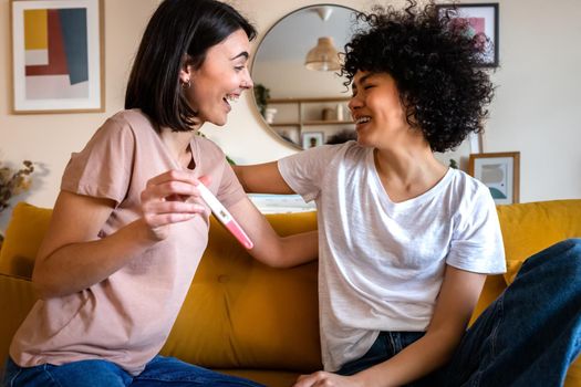 Smiling young woman shows girlfriend positive pregnancy test. Happy multiracial lesbian couple find out pregnancy. LGBTQ family concept.