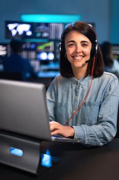 Smiling young female caucasian financial analyst working in the office with laptop and headset looking at camera. Vertical. Stock market and tradning concept.