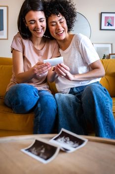 Happy multiracial lesbian pregnant couple looking the first baby ultrasounds at home. Vertical image. LGTBQ love and family concept.