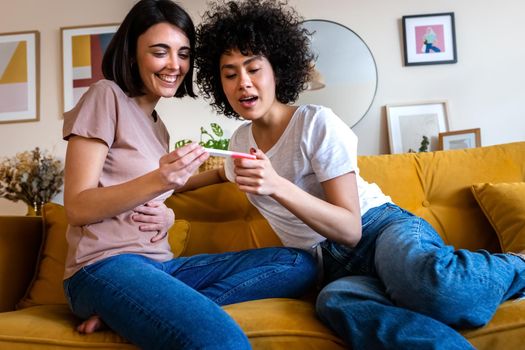 Young multiracial lesbian couple looking at positive pregnancy test together at home living room. LGTBQ love concept.