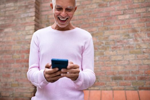 Smiling caucasian adult man looking at cellphone. Browsing through social media. Orange brick wall background. Copy space. Lifestyle and technology concept.