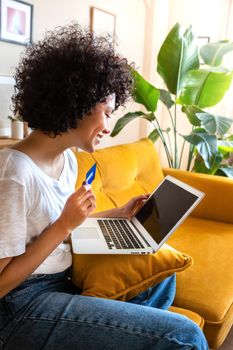 Side view of happy African american woman using credit card and laptop to shop online or order food at home sitting on sofa. Vertical image. Lifestyle and e-commerce concept.