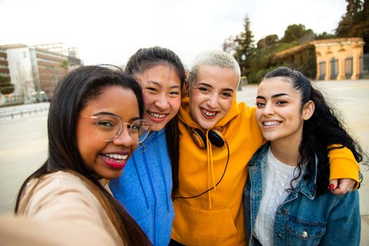 Group of happy and smiling multiracial female teen college student friends take selfie and have fun together outdoors in the city. Gen z friendship and technology concept.