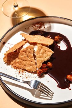 Close-up shot of a gourmet vanilla ice-cream with crunchy biscuits and hazelnuts, drenched in dark chocolate. Served on a white plate with black edging and fork, with a glass of wine. Restaurant cooking concept.