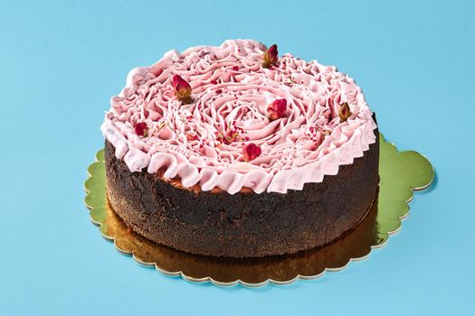 Close-up shot of a pink cream cake decorated with a dried rosebuds, standing on a gold plateau against a blue studio background. Pastries, delicious holiday bake.