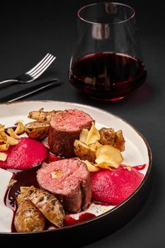 Close-up photo of a tasty roasted veal meat with jerusalem artichokes and sauce. Served on a white plate with black edging and glass of red wine, dark table. Restaurant cooking concept.