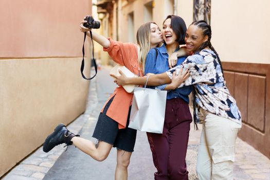 Delightful elegant multiracial women standing against blurred urban building and taking self portrait on camera while rejoicing at purchases