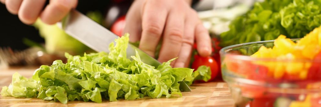 A male chef cuts a green salad with a knife, close-up. Ripe vegetables on the kitchen table for cooking in a restaurant