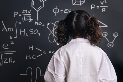 rear view of a little girl science student in lab coat on school blackboard background with hand drawings science formula pattern, back to school and successful female career concept