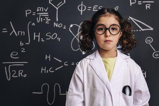 serious little girl science student with glasses in lab coat on school blackboard background with hand drawings science formula pattern, back to school and successful female career concept