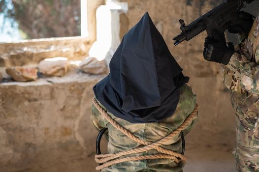 A caucasian woman in an army uniform holds a hostage with a bag on her head at the sight of a machine gun