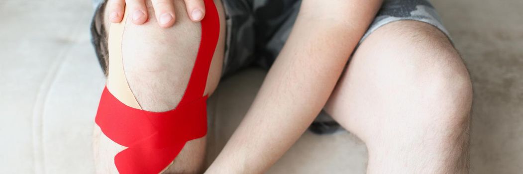 A man sits with a kinesiology tape on his knee, legs close up. Supports knee muscles. Ligament sprain, postoperative rehabilitation