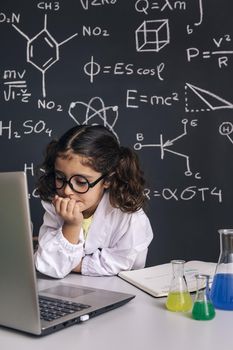 little scientist girl with glasses in lab coat looking to the computer, on school blackboard background with science formulas, back to school and successful female career concept, vertical photo