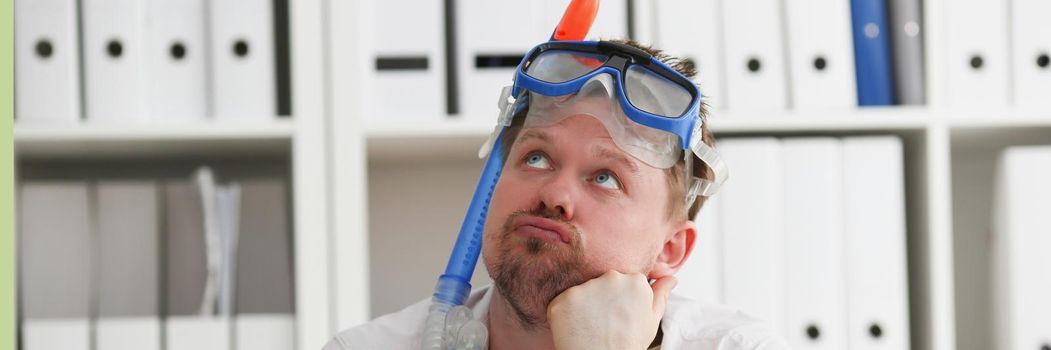 A man wearing a scuba diver's mask is sitting in the office, close-up. Tired employee at work dreams of vacations and diving