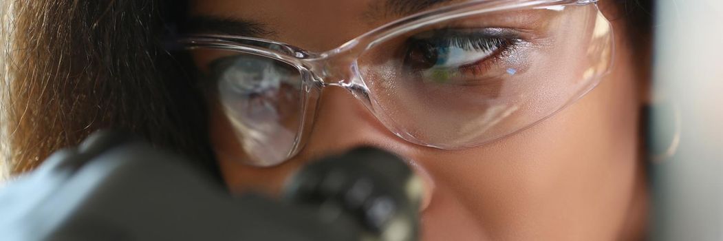 A dark-skinned woman looks through a microscope, a close-up of the face. Scientist is looking for microorganisms to treat a strain of coronavirus