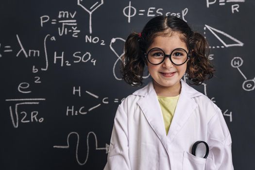 cheerful little girl science student with glasses in lab coat smiles on school blackboard background with hand drawings science formula pattern, back to school and successful female career concept