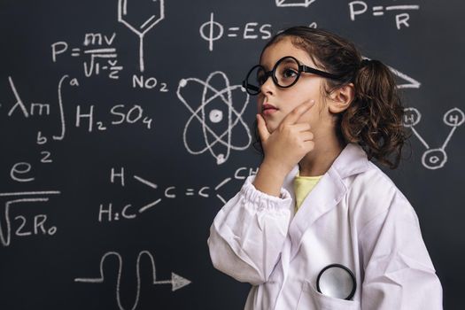 pensive little girl science student in lab coat on school blackboard background with hand drawings science formula pattern, back to school and successful female career concept