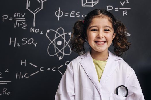 happy little girl science student in lab coat on school blackboard background with hand drawings science formula pattern, back to school and successful female career concept