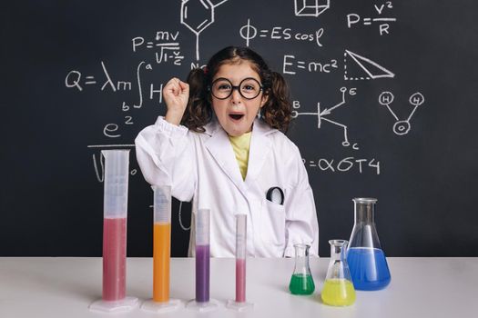 happy scientist child with glasses in lab coat with chemical flasks celebrating an idea, school blackboard background with science formulas, back to school and successful female career concept