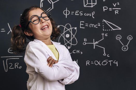 funny little girl science student with glasses in lab coat laugh on school blackboard background with hand drawings science formula pattern, back to school and successful female career concept