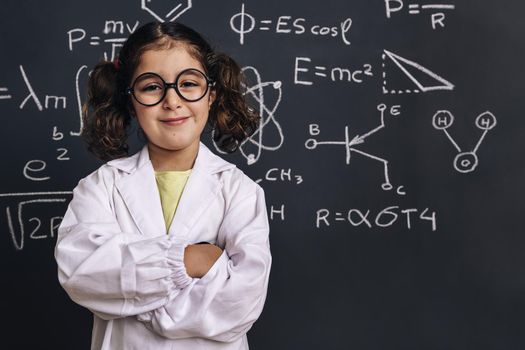 cheerful little girl science student with glasses in lab coat smiles on school blackboard background with hand drawings science formula pattern, back to school and successful female career concept