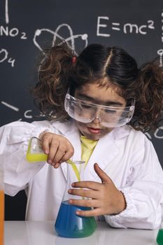 scientist kid with goggles and gloves in lab coat mixing chemical liquids in flasks, blackboard background with science formulas, explosion in the laboratory, back to school concept, vertical photo