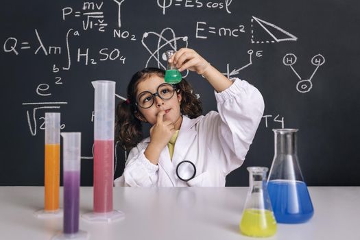 child scientist with glasses in lab coat looking a flask with chemical liquid, on blackboard background with science formulas, concept of back to school and successful female career