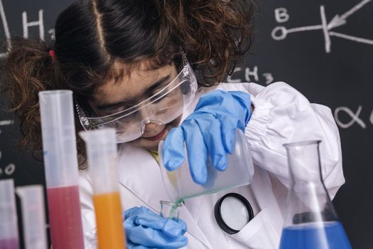 little scientist girl with goggles and gloves in lab coat mixing chemical liquids in flasks, blackboard background with science formulas, explosion in the laboratory, back to school concept