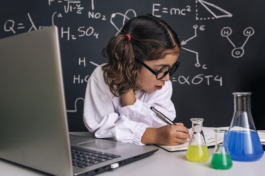 scientist child with glasses studying with a computer and taking notes, on school blackboard background with science formulas, back to school and successful female career concept