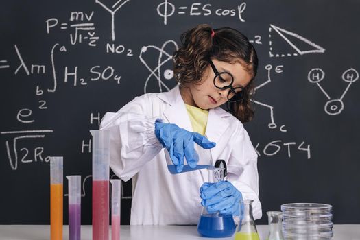 little scientist kid with glasses and gloves in lab coat mixing chemical liquids in flasks, on blackboard background with science formulas, concept of back to school and successful female career