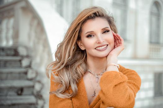 A middle-aged woman looks like a good blonde with curly beautiful hair and makeup on the background of the building. She is wearing a yellow sweater and looking at the camera