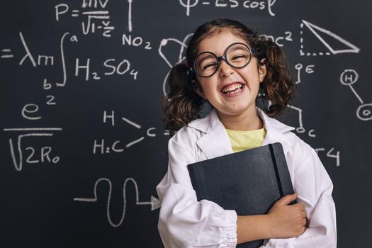 funny little girl science student in lab coat laughing with a notebook on school blackboard background with science formulas, back to school and successful female career concept