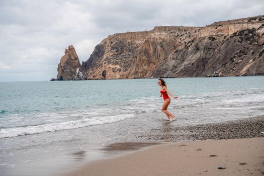 A beautiful and sexy brunette in a red swimsuit on a pebble beach, Running along the shore in the foam of the waves.