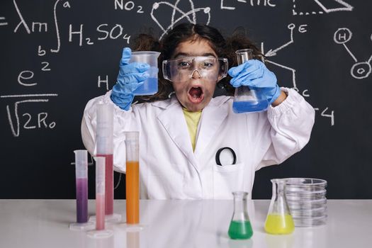funny scientist child with gloves and goggles in lab coat with chemical flasks, blackboard background with science formulas, explosion in the laboratory, back to school concept