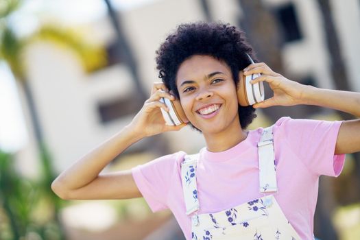 Smiling African American woman in casual clothes walking among trees in urban park listening to music with headphones.