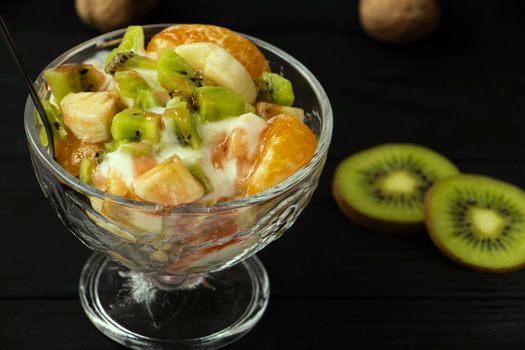 fruit salad in a glass bowl. Kiwi, banana and tangerine with yoghurt and fruit topping. A healthy, vitamin-rich snack. Healthy nutrition concept. Black wood background. High quality photo