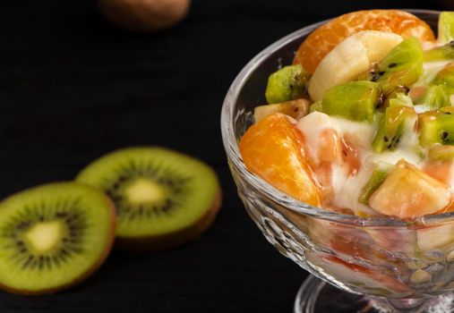 tropical fruit salad in glass bowl. Kiwi, banana and tangerine with yogurt and topping. Veggie dessert. Healthy food concept. Black wood background. High quality photo