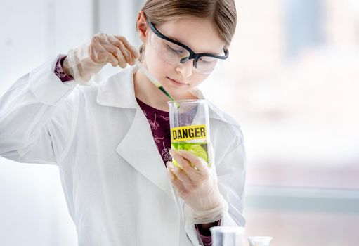 Smart girl doing scientific chemistry experiment wearing protection glasses, and add liquid to bottle with title danger. Schoolgirl with chemical equipment on school lesson