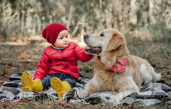 Little girl child with golden retriever dog sitting on the ground on plaid in the forest at autumn. Female kid petting cute doggie pet outdoors