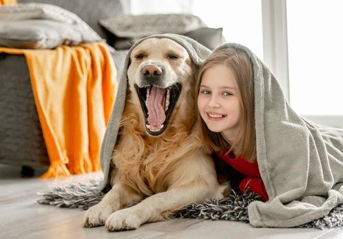 Cheerful child girl lies with golden retriever dog under plaid. Happy preteen girl with purebred doggy labrador spend time on floor.