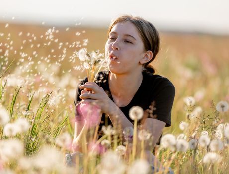 Pretty girl teenager sitting in field and blows dandelions outdoors. Beautiful young female person at nature in sunny day portrait