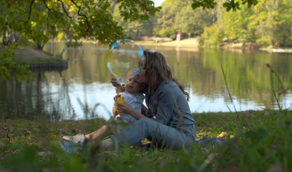 Happy Mother and her little son playing together outdoor, blowing soap bubbles, having fun in the park near the lake. Family, Mother's Day Joy