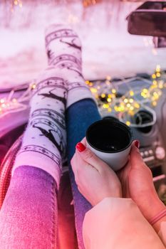 Woman legs with socks hand holding thermos cup of coffee or tea in winter car. Christmas socks. Garland defocused lights. Winter travel.
