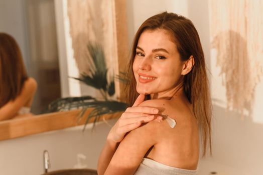 Happy young woman wrapped in towel using nourishing cream or body lotion for her smooth and silky skin after morning shower, rubbing her shoulder, copy space, sun glare. Web banner.